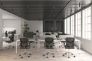 A modern office space features several empty workstations with black Silla Brescia mesh chairs and computer screens. Large windows on the right flood the room with natural light, while a plant is visible in the back corner.